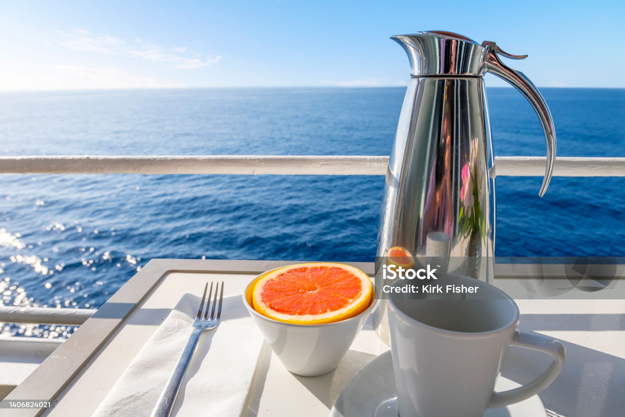 A carafe of coffee with a coffee cup alongside a cut grapefruit on a ocean view balcony of a cruise ship at sea on a summer day.